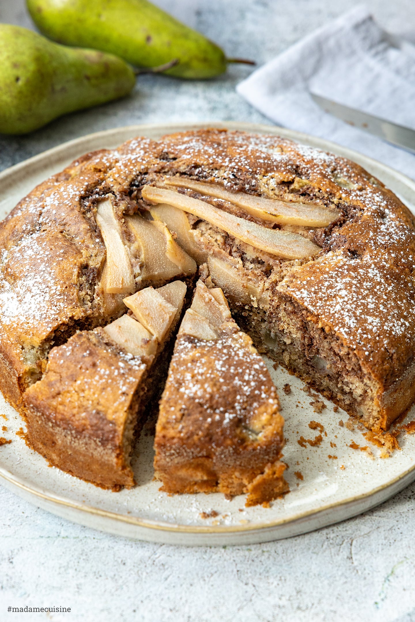 Angeschnittener Kuchen mit Birnenspalten-Topping und ganzen Birnen im Hintergrund