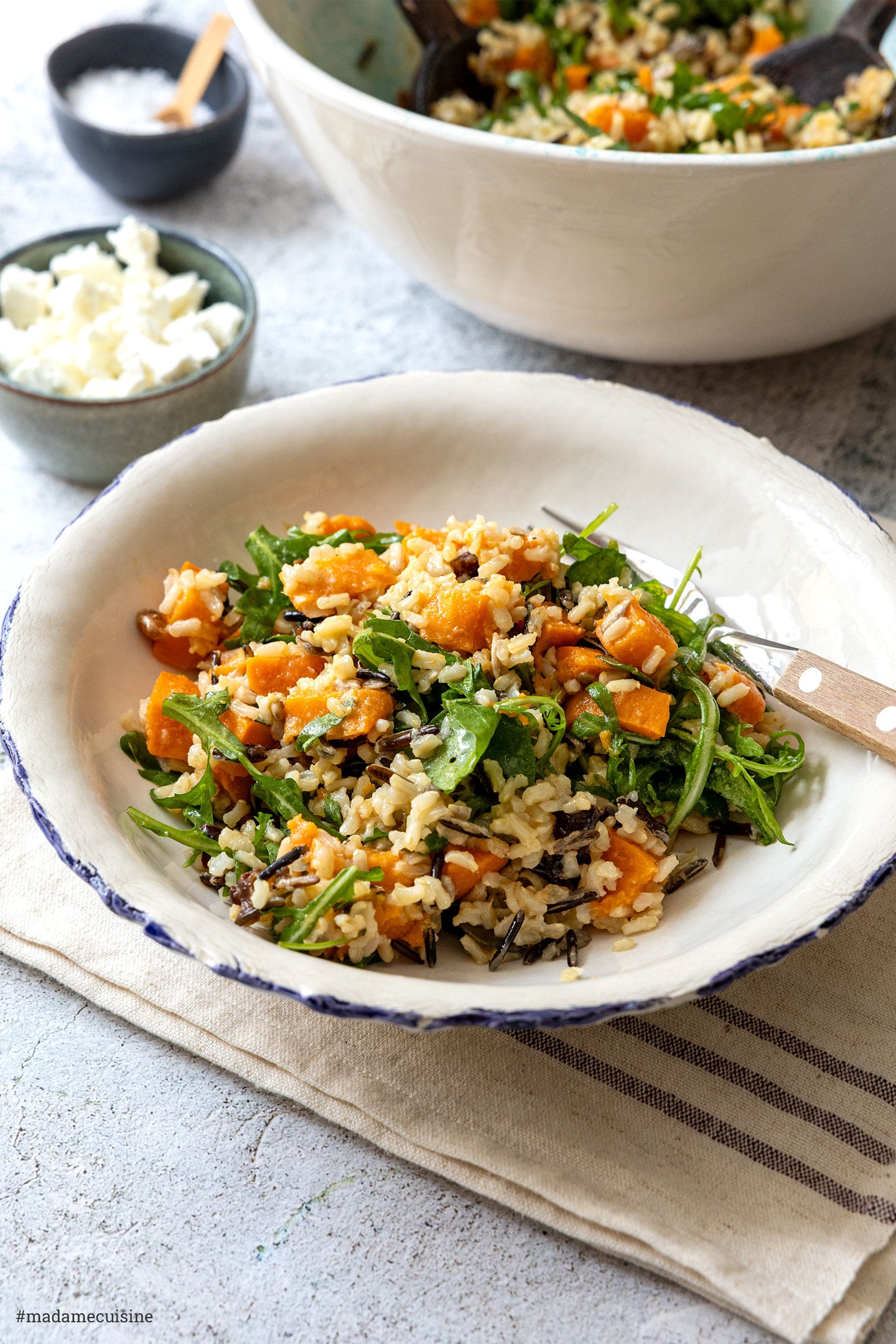 Wildreissalat mit Süßkartoffeln und Rucola auf einem Teller, dahinter eine Servierschüssel und Feta im Schälchen
