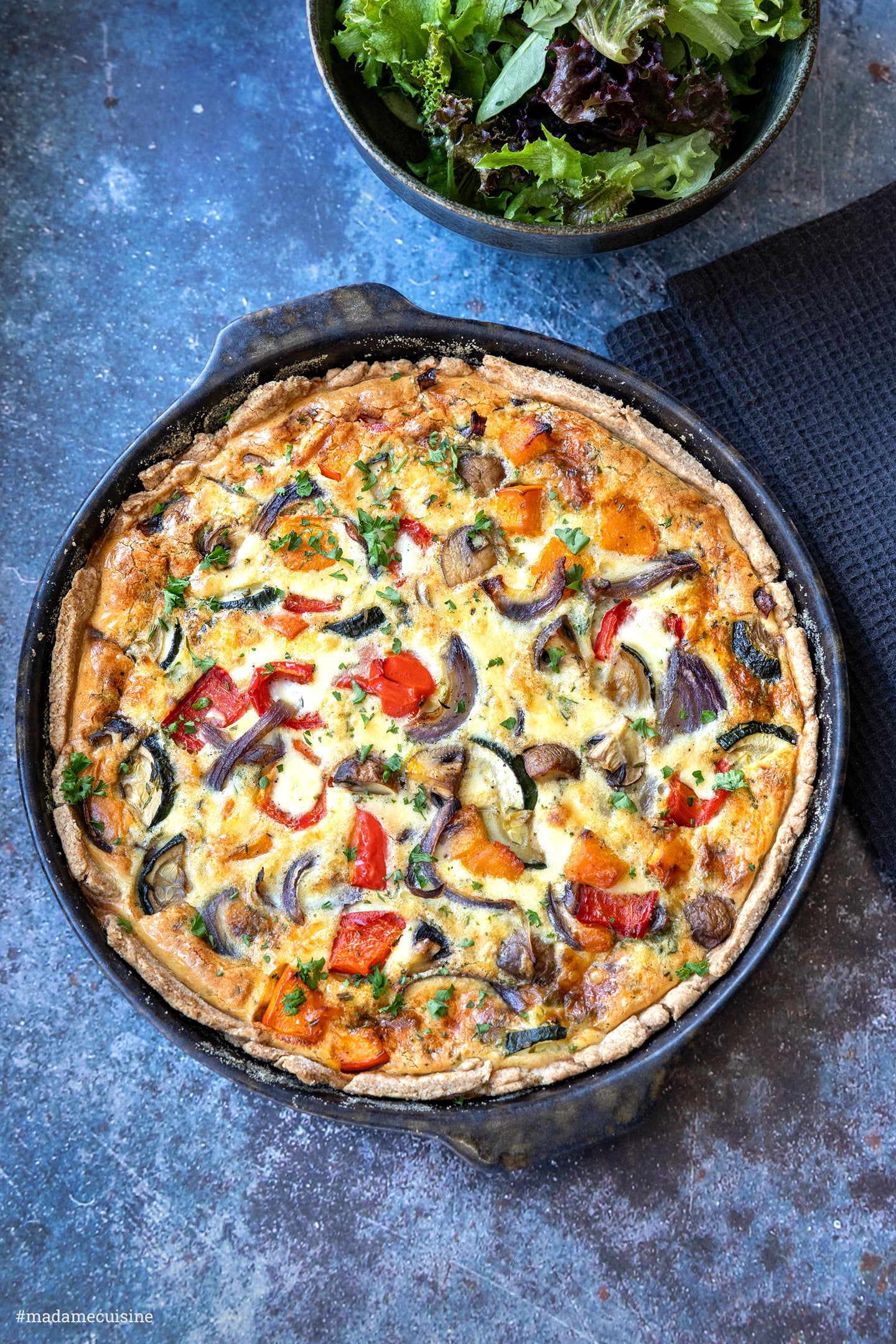 Ganze Kürbis-Quiche von oben in dunkler Form, daneben eine Schale mit grünem Salat
