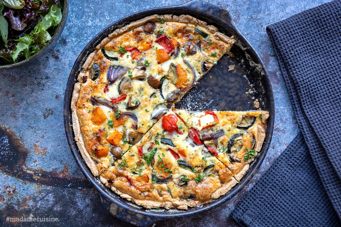 Kürbis-Quiche mit Hokkaido, Paprika und Zucchini in dunkler Quicheform, ein Stück herausgeschnitten