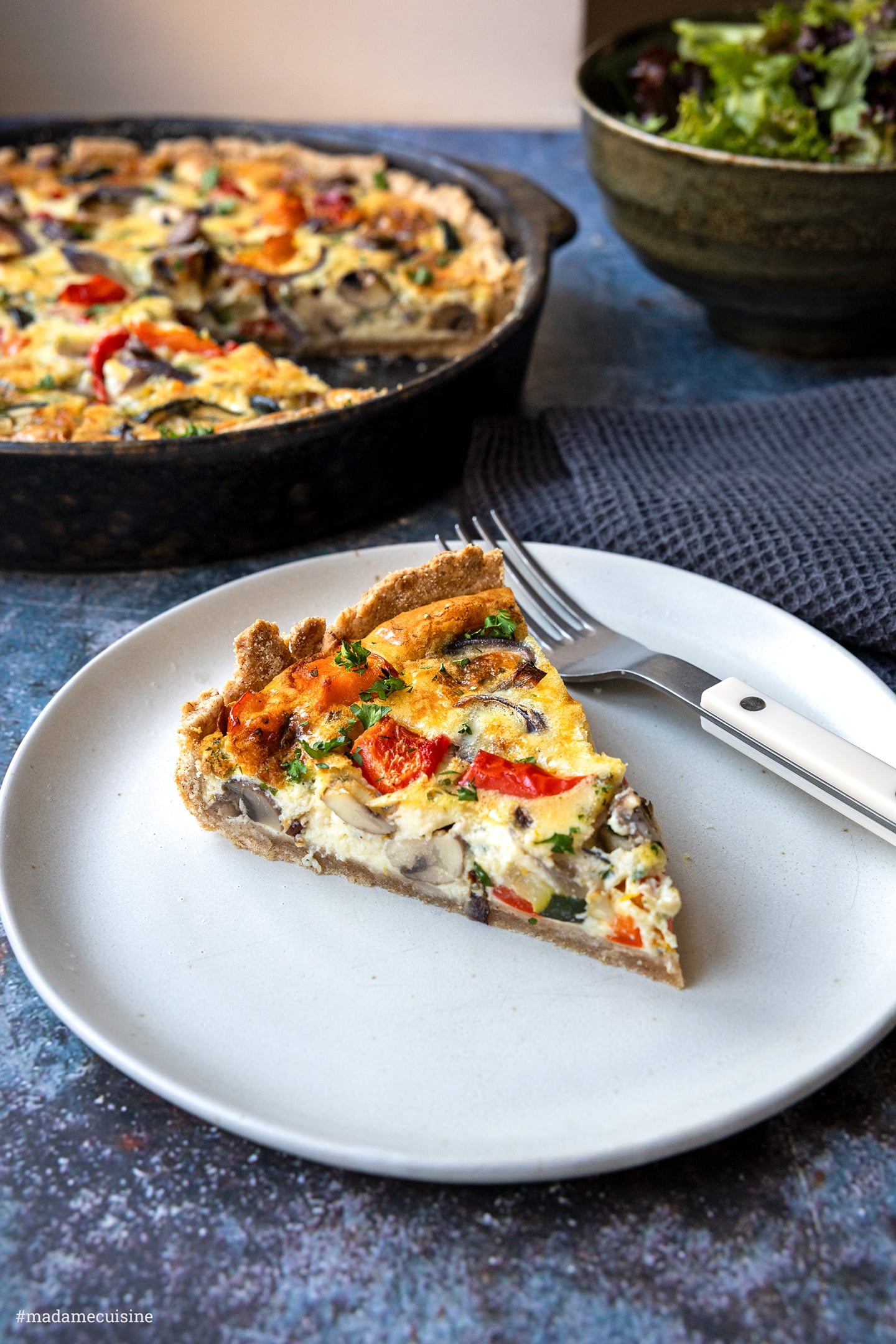 Kürbis-Quiche, ein Stück auf hellem Teller mit Gabel, im Hintergrund die ganze Quiche