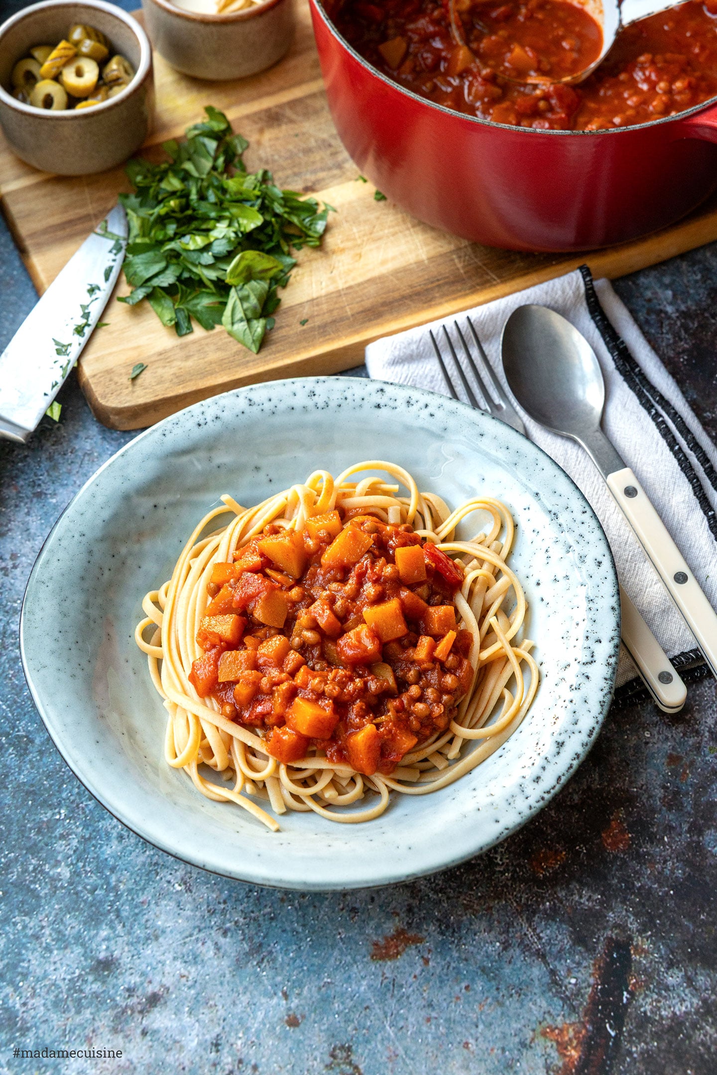 Linguine mit Kürbis-Linsen-Bolognese, dahinter ein Schneidebrett mit gehackten Kräutern und der Topf