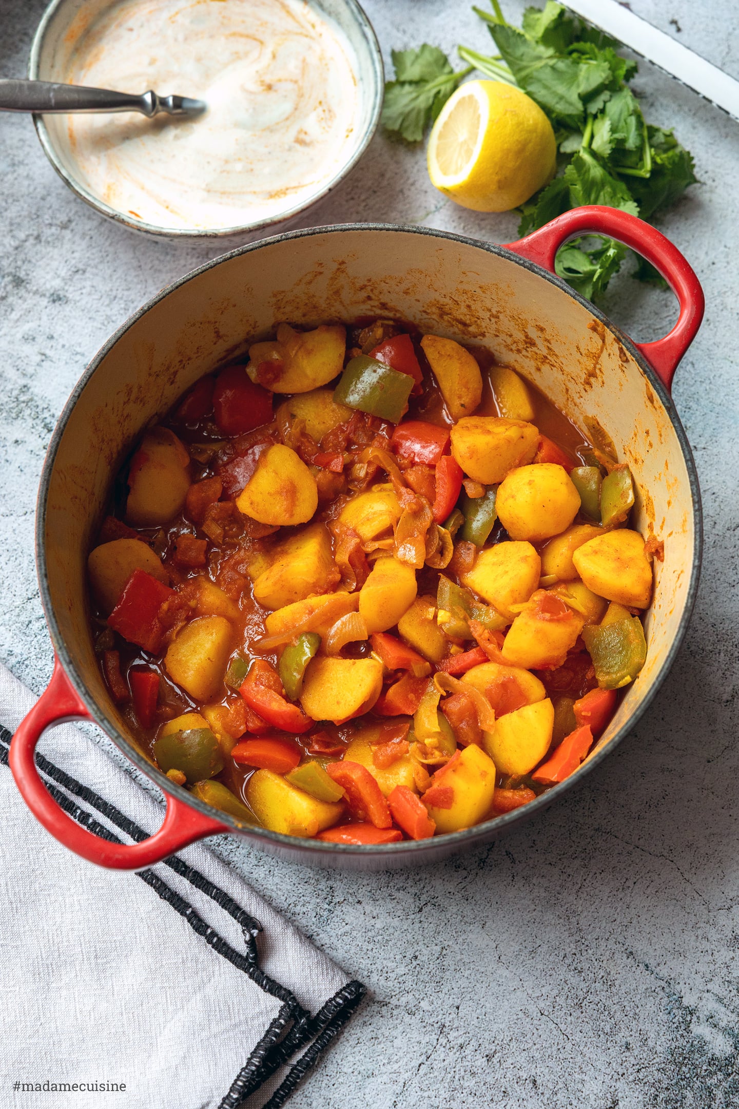 Kartoffeln, Paprika und Zwiebeln in dicker Tomatensauce im roten gusseisernen Topf, dahinter Zitrone und Koriander
