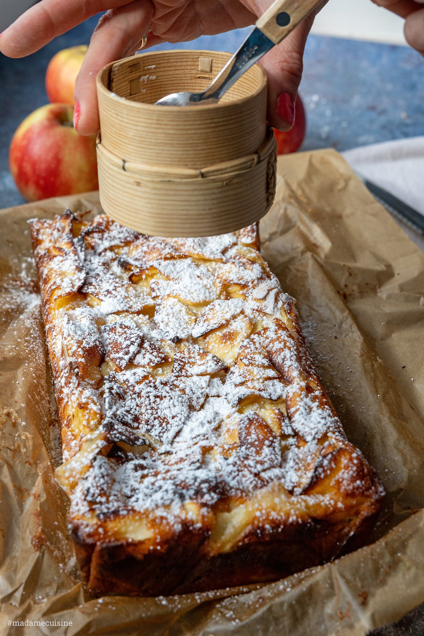 Ganzer Kuchen auf Backpapier wird mit Puderzucker bestäubt, Äpfel im Hintergrund